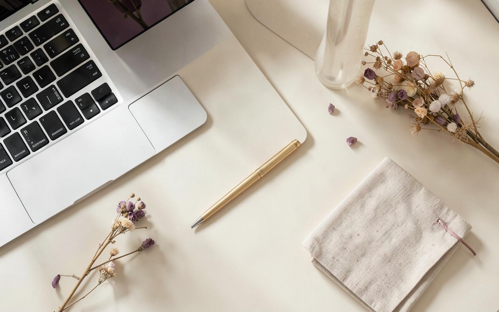 A sunlit ivory desk with a laptop, gold pen and dried florals